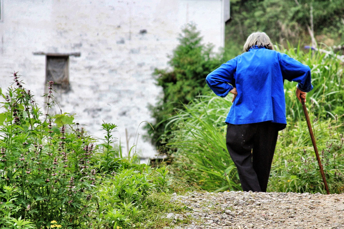 Donna anziana in giacca blu che cammina con un bastone lungo un sentiero